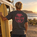 Members of Nowhere Palm Tee in Faded Black, worn at the beach beside a van and surfboard
