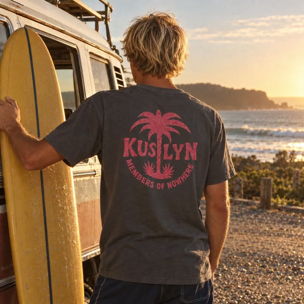Members of Nowhere Palm Tee in Faded Black, worn at the beach beside a van and surfboard